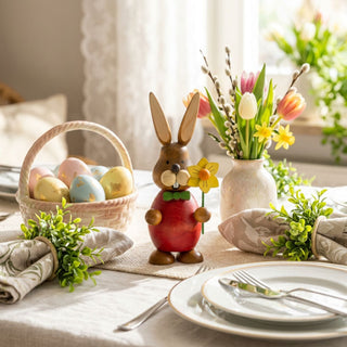 Osterhase mit Blume (ca. 13 cm), farbig gestaltete Figur mit rotem Oberteil und grüner Schleife, hält eine gelbe Blüte. Platziert auf einem gedeckten Tisch mit Ostereiern im Korb, Geschirr und Frühlingsblumen in heller Wohnatmosphäre.