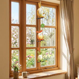 Girlande mit Osterhasen (ca. 49 cm), farbige Hängedeko mit drei Hasenfiguren auf kleinen Holzschaukeln, verbunden durch eine Schnur mit Blumen- und Blattelementen. Aufgehängt vor einem Fenster mit Blick in einen blühenden Frühlingsgarten; auf der Fensterbank stehen Blumentopf und Tasse.
