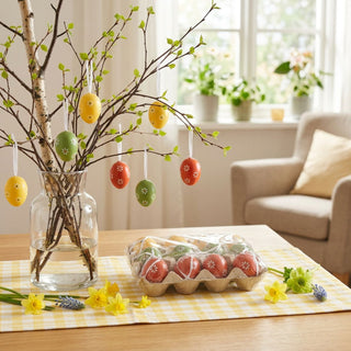 Ostereier zum Aufhängen (12 Stück), farbig bemalte Eier mit weißen Blumenmustern und Aufhängebändern. Dekorativ an Zweigen in einer Glasvase aufgehängt, daneben eine offene Eierbox auf einem Tisch mit Tischläufer und Frühlingsblumen, im Hintergrund ein heller Wohnraum mit Fenster.