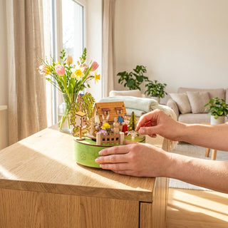 Spieluhr „Osterhasenkarussell“ (ca. 14 cm) mit LED, farbiges Diorama mit Hasen, Haus, Blumen und Zaun auf runder Basis. Steht auf einer Kommode neben Vase mit Frühlingsblumen; eine Hand richtet die Szene, im Hintergrund ein heller Wohnraum mit Sofa.
