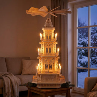Weihnachtspyramide XXL „Gotik“ aus naturbelassenem Holz, ca. 100 cm hoch, elektrisch betrieben und ohne Bestückung, mit mehreren reich verzierten Etagen im gotischen Stil und großem Flügelrad. Warm beleuchtet durch kerzenförmige Lichter, auf einem Tisch vor einem Fenster mit verschneiter Winterlandschaft in einem gemütlichen Wohnraum.