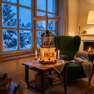 Weihnachtspyramide „Christi Geburt“ aus naturbelassenem Holz mit zwei Etagen und filigranen Figuren, etwa 44 cm hoch, auf einem kleinen Holztisch arrangiert. Mehrere brennende Kerzen treiben das Flügelrad an. Die Szene steht in einem gemütlich eingerichteten Wohnzimmer mit grünem Sessel, Kaminfeuer im Hintergrund und verschneiter Winterlandschaft vor dem Fenster im sanften Abendlicht.