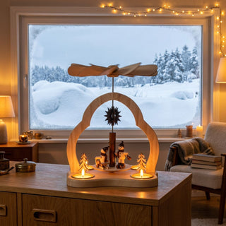 Weihnachtspyramide „Wintersport“ aus naturbelassenem Holz (23 cm) für Teelichter mit Flügelrad. Szene mit Wintersportfiguren und Tannenbäumen unter einem geschwungenen Bogen. Auf einem Tisch vor großem Fenster mit verschneiter Landschaft, warm beleuchtet und dekoriert.