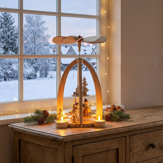 Weihnachtspyramide „Winterwald“ aus naturbelassenem Holz (30 cm) für vier Teelichter mit Flügelrad. Offene Bogenform mit kleinen Winterwald-Figuren und Tannenbäumen. Auf einer Kommode vor verschneitem Fenster, warm beleuchtet und mit Tannenzweigen dekoriert.