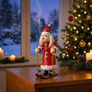 Farbiges Räuchermännchen „Weihnachtsmann auf Ski“ (17 cm) aus Holz mit rotem Mantel, Weihnachtsmütze, langen weißen Haaren und Skiern an den Füßen. Die Figur hält zwei Skistöcke in den Händen, während aus der Öffnung im Mund feiner Rauch aufsteigt. Das Räuchermännchen steht auf einem Holzmöbel vor einem Fenster mit verschneiter Winterlandschaft. Im Hintergrund sorgen ein festlich geschmückter Weihnachtsbaum, Kerzenlicht und Tannenzweige für eine warme und stimmungsvolle Weihnachtsatmosphäre.