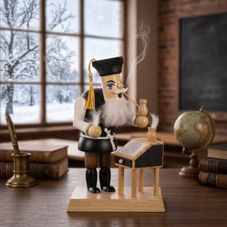 Farbiges Räuchermännchen „Lehrer“ (23 cm) aus Holz mit schwarzem Doktorhut, weißem Bart und Pfeife. Aus der Pfeife steigt feiner Rauch auf. Die Figur steht neben einem kleinen Lehrerpult mit Schreibfeder und aufgeschlagenem Heft. Im Hintergrund sind ein Globus, Bücher, eine Tafel und ein großes Fenster mit verschneiter Winterlandschaft zu sehen. Die detailreiche Szene erinnert an ein historisches Klassenzimmer und vermittelt eine traditionelle, akademische Atmosphäre.