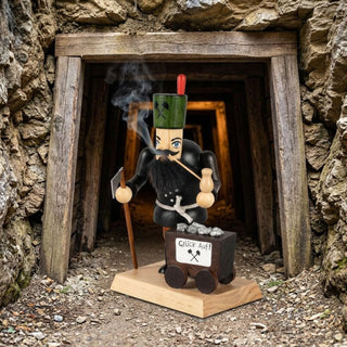 Farbiges Räuchermännchen „Bergmann mit Lore“ (23 cm) aus Holz mit traditioneller Bergmannskleidung, grünem Schachthut, weißem Bart, Pfeife und Grubenwerkzeug. Aus der Pfeife steigt feiner Rauch auf. Neben der Figur steht eine kleine Lore mit Erzbrocken und der Aufschrift „Glück Auf!“. Das Räuchermännchen ist vor dem Eingang eines historischen Bergwerksstollens platziert. Die rustikale Kulisse aus Holzstützen, Felsgestein und Grubengang unterstreicht den traditionellen Bergbaucharakter der Szene.