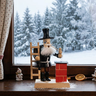 Farbiges Räuchermännchen „Schornsteinfeger“ (23 cm) aus Holz mit schwarzer Schornsteinfegerkleidung, Zylinder, weißem Bart, Pfeife sowie einer Holzleiter und einem Kehrbesen. Aus der Pfeife steigt feiner Rauch auf. Neben der Figur steht ein roter Schornstein, aus dem ebenfalls Rauch austritt. Das Räuchermännchen ist auf einer Fensterbank vor einem großen Fenster mit verschneiter Winterlandschaft platziert. 