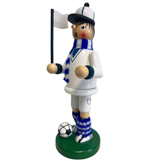 Räuchermännchen „Fußballer“ aus Holz, ca. 23 cm groß, in weiß-blauer Farbgestaltung aus schräger Seitenansicht. Die Figur steht auf einem runden grünen Sockel neben einem schwarz-weißen Fußball, trägt ein weißes Outfit mit blauen Details, einen blau-weiß gestreiften Schal sowie gestrickte Beinwärmer und hält eine kleine weiße Fahne an einem schwarzen Stab.
