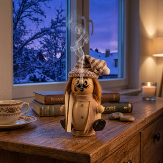 Naturfarbenes Räuchermännchen Wichtel „Skifahrer“ (17 cm) aus Holz mit gestrickter Wintermütze, Schal und einem Paar Skier in der Hand. Aus der Pfeife steigt feiner Rauch auf. Die Figur sitzt auf einer Holzkommode vor einem Fenster mit verschneiter Winterlandschaft. Daneben stehen eine Tasse, Bücher und eine Kerze. Die warme Beleuchtung im Raum und die winterliche Abendstimmung im Hintergrund schaffen eine gemütliche und einladende Atmosphäre.