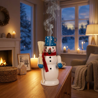 Farbiges Räuchermännchen „Schneemann mit Besen“ (10 cm) aus Holz. Die Schneemannfigur trägt einen blauen Hut, einen roten Schal und blaue Handschuhe und hält einen Besen in der Hand. Aus dem geöffneten Mund steigt feiner Räucherrauch auf. Das Räuchermännchen steht auf einem Holztisch in einem gemütlich beleuchteten Wohnzimmer mit Kaminfeuer, Kerzen und einer verschneiten Winterlandschaft vor dem Fenster im Hintergrund.