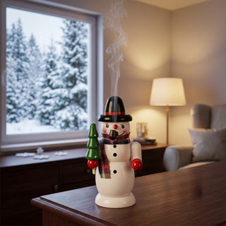 Farbiges Räuchermännchen „Schneemann mit Tanne“ (10 cm) aus Holz. Die Schneemannfigur trägt einen schwarzen Hut mit rotem Band, einen karierten Schal und rote Handschuhe. In einer Hand hält sie eine grüne Tanne, aus dem geöffneten Mund steigt feiner Rauch auf. Das Räuchermännchen steht auf einem Holzmöbel in einem gemütlichen Wohnraum. Im Hintergrund sind ein Fenster mit verschneiter Winterlandschaft, eine Stehlampe und eine Sitzecke zu sehen.Geben Sie Ihr Feedback zu BizChat ab.