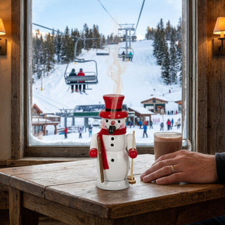 Farbiges Räuchermännchen „Schneemann mit Ski“ (10 cm) aus Holz. Die kleine Schneemannfigur trägt einen roten Zylinderhut, einen roten Schal sowie rote Handschuhe und hält ein Paar Ski und Skistöcke. Aus dem geöffneten Mund steigt feiner Rauch auf. Das Räuchermännchen steht auf einem Holztisch vor einem Fenster mit Blick auf ein verschneites Skigebiet mit Sessellift und Wintersportlern. Daneben stehen eine Tasse Heißgetränk und eine Hand, die die kompakte Größe der Figur verdeutlicht.