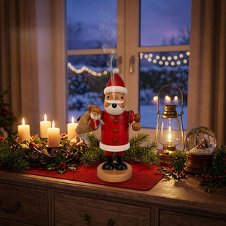 Farbiges Räuchermännchen „Weihnachtsmann“ (21 cm) aus Holz mit rotem Mantel, roter Mütze und weißem Bart. Die Figur hält einen Geschenkesack und eine Pfeife in den Händen, aus deren Mundbereich Rauch aufsteigt. Das Räuchermännchen steht auf einer festlich dekorierten Kommode mit Kerzen, Tannenzweigen, Laterne und Schneekugel. Im Hintergrund ist ein Fenster mit winterlicher Abendstimmung und warmen Lichterketten zu sehen.
