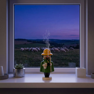 Farbiges Räuchermännchen Mini „Schäfer“ (9 cm) aus Holz mit grünem Mantel, gelbem Hut, Pfeife und Hirtenstab. Aus der Pfeife steigt feiner Rauch auf. Die Figur hält ein kleines Schaf im Arm und steht auf einer Fensterbank vor einem großen Fenster. Im Hintergrund ist eine Schafherde auf einer Weide in der Abenddämmerung zu sehen. Die ruhige Landschaft und die dezente Inneneinrichtung mit Kerzen und Pflanze schaffen eine idyllische und beschauliche Atmosphäre.