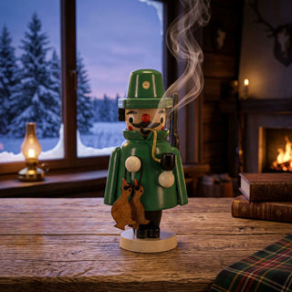Farbiges Räuchermännchen Mini „Jäger“ (9 cm) aus Holz mit grünem Jagdmantel, grünem Hut, Pfeife und geschultertem Gewehr. Aus der Pfeife steigt feiner Rauch auf. Die Figur trägt zwei erlegte Hasen und steht auf einem rustikalen Holztisch in einer gemütlichen Jagdhütte. Im Hintergrund sind ein Kaminfeuer, Bücher, eine Öllampe und ein Fenster mit verschneiter Winterlandschaft zu sehen. Die warme Beleuchtung und die winterliche Kulisse schaffen eine traditionelle und behagliche Atmosphäre.