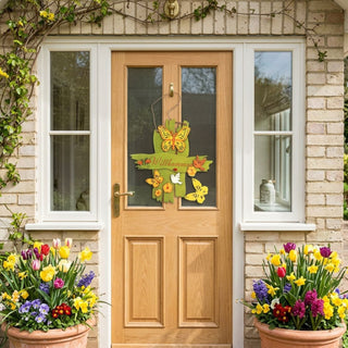 Dekoschild „Willkommen“ (ca. 29 cm), farbiges Hängeschild mit Schriftzug und Schmetterlingen sowie Blumenmotiven, an einer Haustür befestigt. Umgeben von blühenden Pflanzen in großen Töpfen vor einem gepflegten Hauseingang.