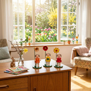 Blumenkinder Mädchen (3er-Set, je ca. 11 cm), drei farbig gestaltete Figuren mit Blumen in den Händen auf grünen Sockeln. Platziert auf einer Holzkommode neben Vase mit Zweigen und Osterhase, vor einem großen Fenster mit Blick in einen blühenden Frühlingsgarten.