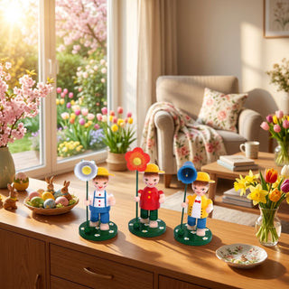 Blumenkinder Jungen (3er-Set, je ca. 11 cm), drei farbig gestaltete Figuren mit Hüten und Blumen in den Händen auf grünen Sockeln. Platziert auf einer Holzkommode mit Osterdeko, Blumen und Ostereiern, vor einem großen Fenster mit Blick in einen blühenden Frühlingsgarten.