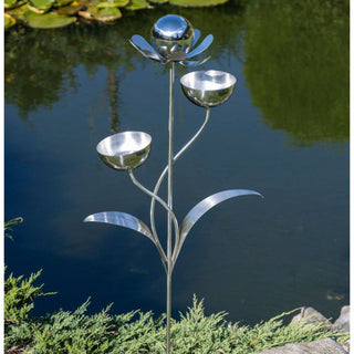 Vogeltränke „Blume“ aus Edelstahl, 116 cm hoch, mit zwei runden Tränkschalen an geschwungenen Stielen und dekorativer Blüte an der Spitze, befestigt mit einem stabilen Erdspieß, aufgestellt im Garten am Teichufer mit Wasser, Pflanzen und Steinen im Hintergrund, geeignet als elegante Vogeltränke und Gartendekoration.