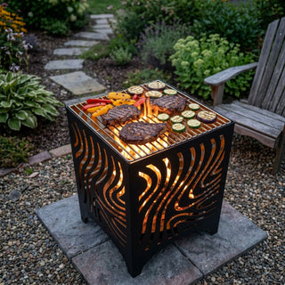Feuerwürfel mit Grillfunktion (ca. 50 cm hoch) aus schwarzem Metall mit dekorativen, geschwungenen Ausschnitten, durch die das Feuer im Inneren sichtbar lodert. Auf dem Grillrost liegen mehrere Steaks sowie gegrilltes Gemüse wie Zucchini und Paprika. Der Feuerwürfel steht auf einer Steinplatte in einem gepflegten Garten mit Kiesboden, Gartenweg, Pflanzen und einem Holzstuhl im Hintergrund.