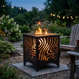 Feuerwürfel mit Grillfunktion (ca. 50 cm hoch) aus schwarzem Metall mit dekorativen, geschwungenen Ausschnitten, durch die das Feuer im Inneren sichtbar lodert. Auf der Oberseite befindet sich ein Grillrost. Der Feuerwürfel steht auf einer Steinplatte in einem gemütlichen Garten bei Abenddämmerung, umgeben von Kies, blühenden Pflanzen, einem Gartenstuhl und einer stimmungsvollen Lichterkette im Hintergrund.