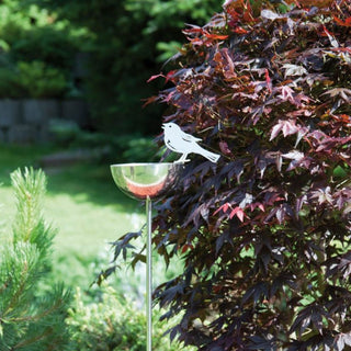 Edelstahl‑Vogeltränke als Gartenstecker mit rundem Wasserschälchen und dekorativem Vogelmotiv, freistehend im grünen Garten neben einem roten Zierstrauch – moderne und wetterfeste Gartendeko für Vögel.