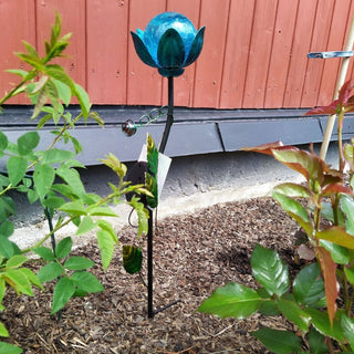 LED Solar‑Gartenstecker „Blume“ in Blau mit leuchtender Glasblüte und Metallblättern, solarbetrieben, dekorativ in einem Gartenbeet zwischen grünen Pflanzen platziert.