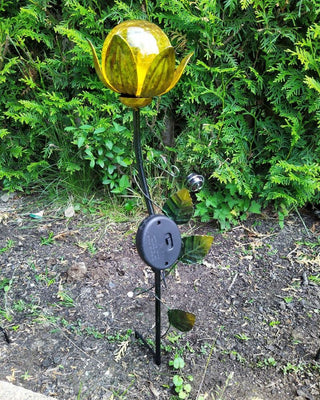Solarbetriebener Gartenstecker in Blumenform mit gelber Glaskugel, aufgestellt im Beet vor grünem Buschwerk.