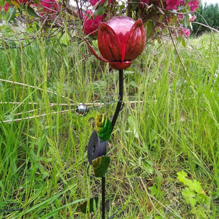 Solarbetriebener Gartenstecker in roter Blumenform mit Glaskugel, aufgestellt im hohen Gras vor blühenden Sträuchern.