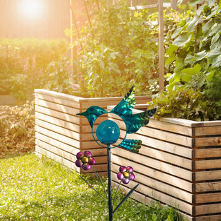 LED Solar‑Gartenstecker „Vogel“ in Blau aus Metall und Glas mit integrierter LED‑Glaskugel und Blumenapplikationen, solarbetrieben, dekorativ im Garten vor Hochbeeten platziert.