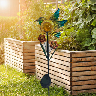 LED Solar‑Gartenstecker „Vogel“ aus Metall und Glas mit integrierter gelber LED‑Glaskugel und Blumenapplikationen, solarbetrieben, dekorativ im Garten vor Holz‑Hochbeeten platziert.