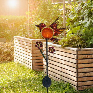 LED Solar‑Gartenstecker „Vogel“ in Orange aus Metall und Glas mit orangefarbener LED‑Glaskugel und dekorativen Blumenapplikationen, solarbetrieben, im Garten vor Holz‑Hochbeeten platziert.