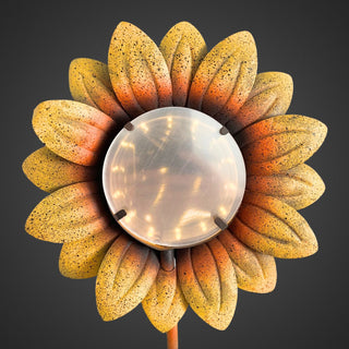 Detailansicht eines Gartensteckers in Sonnenblumenform. Gelb‑orange Blütenblätter umgeben eine runde, helle Mitte mit sichtbarem Tunnellichteffekt und integrierter Solar‑LED. Oberfläche der Blütenblätter leicht strukturiert, vor dunklem Hintergrund.