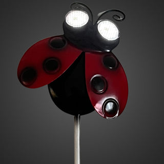 Nahaufnahme eines Gartensteckers „Marienkäfer“ mit rotem Körper und schwarzen Punkten, leuchtende Solar‑LED‑Augen mit wechselnder Blickrichtung, 78 cm hoch, vor dunklem Hintergrund.
