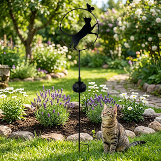 Gartenstecker „Katze stehend“ aus Metall (ca. 84 cm hoch) mit integriertem Solar-LED-Modul. Die schwarze Katzen-Silhouette ist in einen runden Ring mit dekorativem Schmetterling integriert und auf einem stabilen Stab befestigt. Der Gartenstecker steht in einem gepflegten Blumenbeet mit Lavendel und weißen Blüten, umgeben von grünem Rasen, Steinen und einem geschwungenen Gartenweg im sonnigen Hintergrund. Im Vordergrund sitzt zusätzlich eine echte Katze neben dem Gartenstecker.