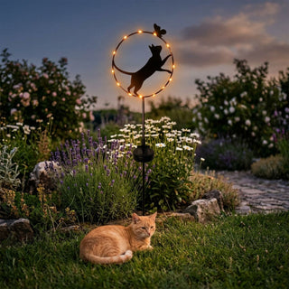 Gartenstecker in Form einer stehenden Katze aus Metall (ca. 84 cm hoch) mit integrierter Solar-LED-Beleuchtung. Die Katzen-Silhouette ist von einem runden Ring mit kleinen warmweiß leuchtenden Lichtern umgeben, auf dessen Rand ein Schmetterling sitzt. Der Gartenstecker steht in einem blühenden Gartenbeet mit Lavendel und weißen Blumen bei Abenddämmerung. Im Vordergrund liegt zusätzlich eine echte Katze im Gras, während im Hintergrund ein Gartenweg, Sträucher und der Abendhimmel zu sehen sind.