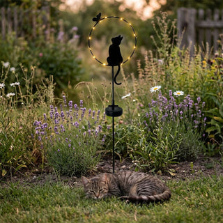 Gartenstecker „Katze sitzend“ aus Metall (ca. 84 cm hoch) mit integriertem Solar-LED-Modul. Die schwarze Silhouette einer sitzenden Katze ist in einen runden Ring mit kleinen warmweiß leuchtenden LEDs und einem dekorativen Schmetterling am oberen Rand integriert. Der Gartenstecker steht in einem naturnahen Blumenbeet mit Lavendel, weißen Blüten und Wildpflanzen bei Abenddämmerung. Im Vordergrund liegt zusätzlich eine echte Katze im Gras.