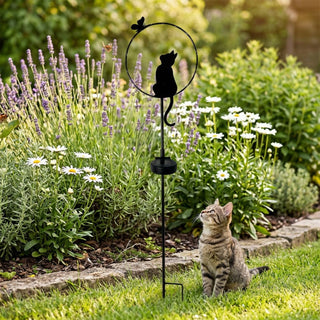 Gartenstecker „Katze sitzend“ aus Metall (ca. 84 cm hoch) mit integriertem Solar-LED-Modul. Die schwarze Silhouette einer sitzenden Katze ist in einen runden Ring mit dekorativem Schmetterling integriert und auf einem schlanken Stab befestigt. Der Gartenstecker steht in einem gepflegten Blumenbeet mit Lavendel und weißen Blüten, umgeben von grünem Rasen und einer niedrigen Steinkante. Im Vordergrund sitzt zusätzlich eine echte Katze im Gras und blickt nach oben.