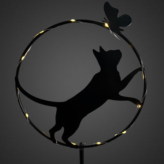 Detailansicht eines Gartensteckers aus schwarzem Metall mit stehender Katzen‑Silhouette in einem runden Rahmen. Die Katze richtet sich zu einem kleinen Schmetterling am oberen Rand des Kreises. Ein feiner Solar‑LED‑Lichtdraht mit warmweißem Licht verläuft entlang des Rahmens und hebt die Konturen hervor. Gesamthöhe des Gartensteckers 84 cm.