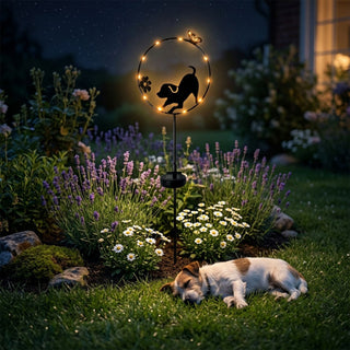 Gartenstecker „Hund spielend“ aus Metall (ca. 84 cm hoch) mit integriertem Solar-LED-Modul. Die Silhouette eines spielenden Hundes ist in einen runden Ring mit kleinen warmweiß leuchtenden LEDs sowie dekorativen Blumen- und Schmetterlingselementen integriert. Der Gartenstecker steht in einem blühenden Gartenbeet mit Lavendel und weißen Blumen bei Nacht. Im Vordergrund liegt zusätzlich ein echter Hund im Gras, während im Hintergrund Gartenpflanzen und ein beleuchtetes Haus zu sehen sind.