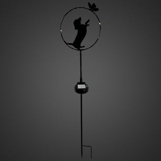 Gartenstecker aus schwarzem Metall mit stehender Hunde‑Silhouette in einem runden Rahmen. Der Hund ist auf den Hinterbeinen dargestellt und greift nach einem kleinen Schmetterling am oberen Rand des Kreises. Ein integrierter Solar‑LED‑Lichtdraht mit warmweißem Licht verläuft entlang des Rahmens. Am Metallstab befindet sich das Solar‑Modul, freistehend vor dunklem Hintergrund.