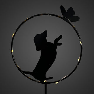 Detailansicht eines Gartensteckers aus schwarzem Metall mit stehender Hunde‑Silhouette in einem runden Rahmen. Der Hund ist auf den Hinterbeinen dargestellt und streckt die Vorderpfoten nach oben zu einem kleinen Schmetterling am Rand des Kreises. Entlang des Rahmens verläuft ein feiner Solar‑LED‑Lichtdraht mit warmweißem Licht, der die Konturen dezent beleuchtet.