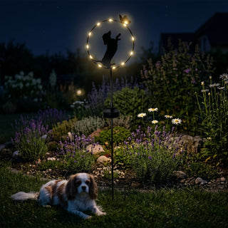 Gartenstecker „Hund stehend“ aus Metall (ca. 84 cm hoch) mit integriertem Solar-LED-Modul. Die Silhouette eines stehenden Hundes ist in einen runden Ring mit kleinen warmweiß leuchtenden LEDs und einem dekorativen Schmetterling integriert. Der Gartenstecker steht in einem blühenden Gartenbeet mit Lavendel und weißen Blumen bei Nacht. Im Vordergrund liegt zusätzlich ein echter Hund im Gras, während im Hintergrund Pflanzen, Steine und ein Haus im Dunkeln erkennbar sind.