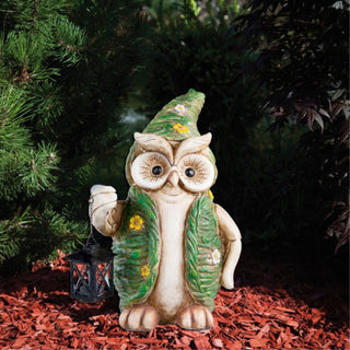 Dekorative Gartenfigur Eule aus Kunststein mit grüner Weste und Hut, steht im Garten auf Rindenmulch und hält eine kleine schwarze Laterne mit LED‑Solar‑Licht, umgeben von Pflanzen.
