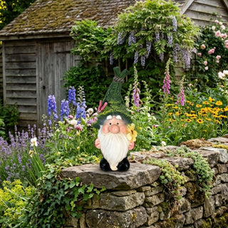 Gartenfigur „Wichtel mit Blume“ (ca. 17 cm hoch) mit langem weißen Bart, runder Nase und grüner, strukturierter Zipfelmütze, die mit einem rosa Schmetterling verziert ist. Der Wichtel hält eine gelbe Blume in der Hand und steht auf einem Kiesweg zwischen Steinplatten in einem gepflegten Garten. Umgeben ist die Figur von Lavendel, bunten Blumen, Farnen und weiteren Zierpflanzen. Im Hintergrund befinden sich Hochbeete mit Gemüse und ein Gartenhaus aus Holz, das von blühenden Pflanzen eingerahmt ist.
Geben Sie