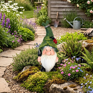 Gartenfigur „Wichtel mit Blume“ (ca. 17 cm hoch) mit langem weißen Bart, runder Nase und grüner, strukturierter Zipfelmütze, auf der ein roter Marienkäfer sitzt. Der Wichtel hält eine kleine schwarze Gartenschaufel und steht zwischen Moos, Steinen und niedrigen Pflanzen in einem liebevoll gestalteten Gartenbeet. Umgeben ist die Figur von bunten Blumen, Lavendel sowie einem geschwungenen Gartenweg aus Steinplatten und Kies. Im Hintergrund sind ein Holzschuppen, eine Gießkanne, Gartenwerkzeug und ein Beet mit