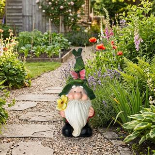 Gartenfigur „Wichtel mit Blume“ (ca. 32 cm hoch) mit langem weißen Bart, runder Nase und grüner, strukturierter Zipfelmütze, die mit einem rosa Schmetterling verziert ist. Der Wichtel hält eine gelbe Blume in der Hand und steht auf einem Kiesweg mit eingelassenen Steinplatten. Die Figur ist umgeben von einem üppig bepflanzten Garten mit Lavendel, Farnen, bunten Blumen und Zierpflanzen. Im Hintergrund sind Hochbeete mit Gemüse sowie ein teilweise von Pflanzen umgebenes Gartenhaus zu sehen.
