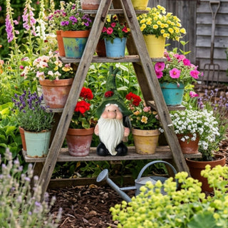 Gartenfigur „Wichtel mit Schaufel“ (ca. 32 cm hoch) mit langem weißen Bart, runder Nase und grüner Zipfelmütze. Der Wichtel hält eine kleine Gartenschaufel und steht auf einer Holzstufe eines dekorativen Pflanzenregals in Form einer Leiter. Das Regal ist dicht mit bunten Blumentöpfen und blühenden Pflanzen bestückt. Im Vordergrund stehen eine Gießkanne und weitere Gartenpflanzen, während im Hintergrund ein üppig bepflanzter Garten mit verschiedenen Blumen zu sehen ist.