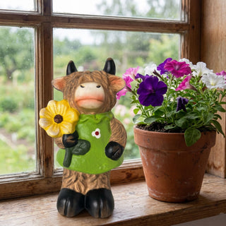 Dekofigur „Galloway-Mädchen mit Blume“ (ca. 14 cm hoch) aus Terrakotta, gestaltet als kleine Kuhfigur mit zotteliger Fellstruktur, schwarzen Hufen und Hörnern. Die Figur trägt ein grünes Kleid mit Blumenmotiv und hält eine gelbe Blüte in der Hand. Sie steht auf einer hölzernen Fensterbank neben einem Blumentopf mit blühenden Petunien in Weiß, Pink und Lila. Im Hintergrund ist ein Fenster mit Blick in einen grünen Garten zu sehen.